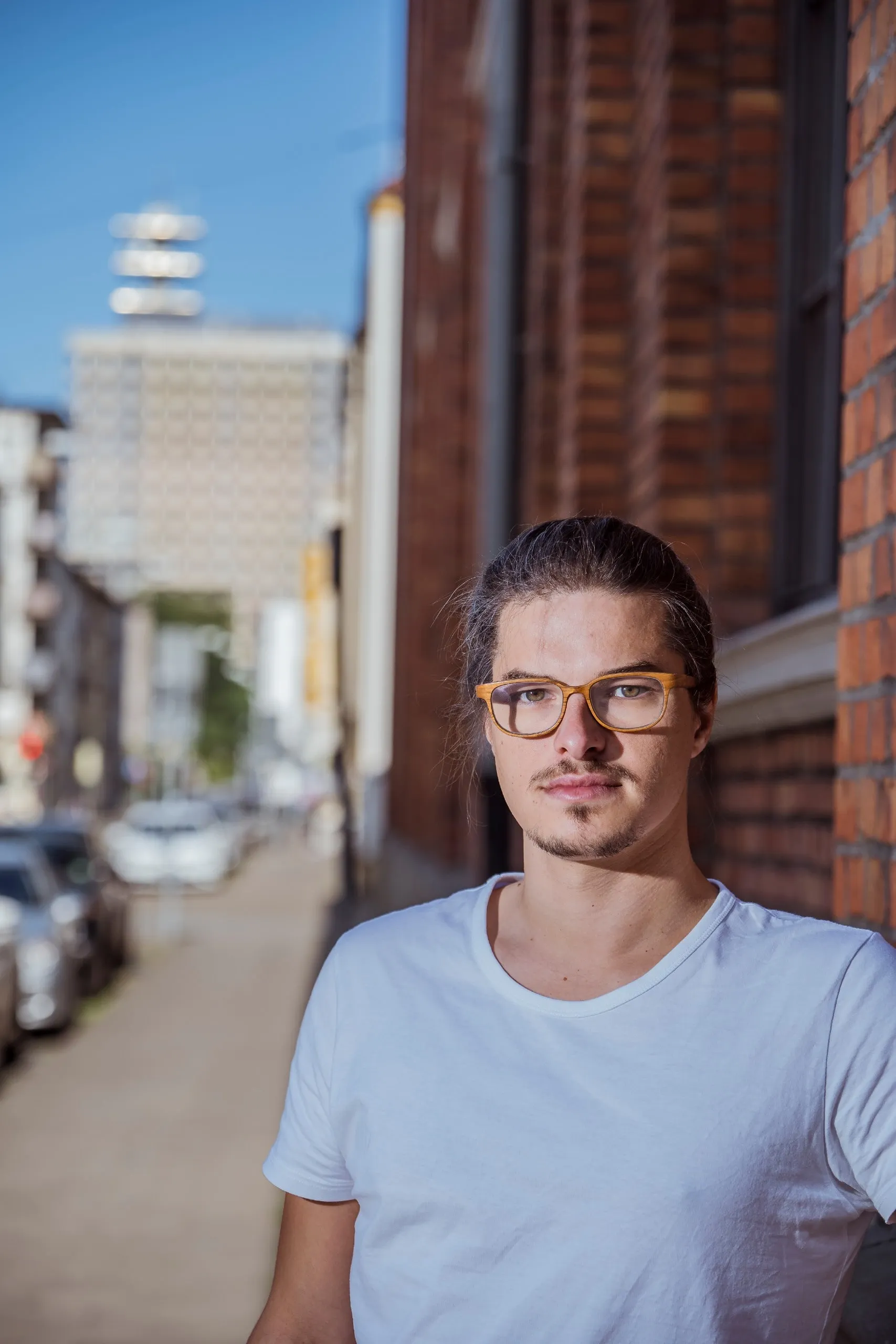 Portrait of Arved Cornelsen wearing glasses and a white T-shirt in front of a brick wall
